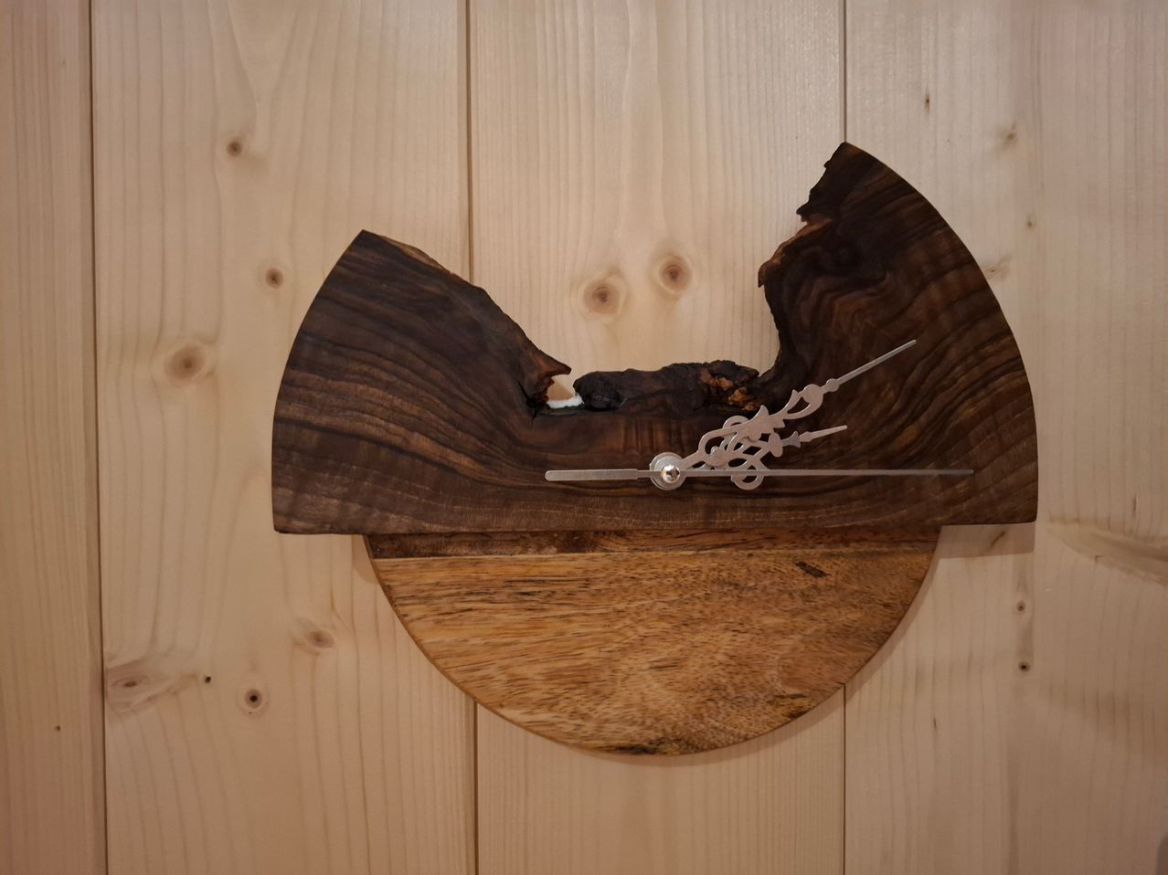 Eine einzigartige Holzuhr aus einem Stück Holz mit einer silberfarbenen Metallhand ist an einer Holzwand montiert.
