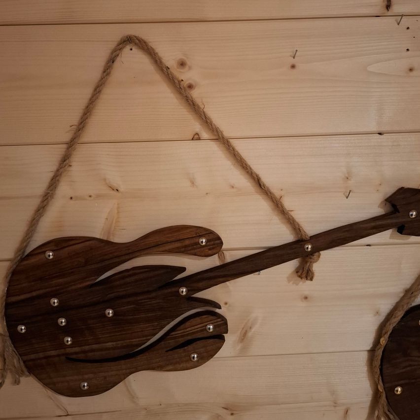 Eine hölzerne Gitarrendekoration mit silbernen Nieten hängt an einer Holzwand, gesichert durch ein Seil.