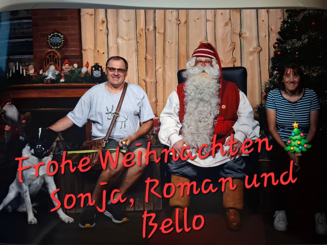 Zwei Männer und eine Frau sitzen mit dem Weihnachtsmann in einem weihnachtlichen Ambiente. Der Weihnachtsmann trägt ein traditionelles rot-weißes Outfit und der Mann links hält einen Hund.