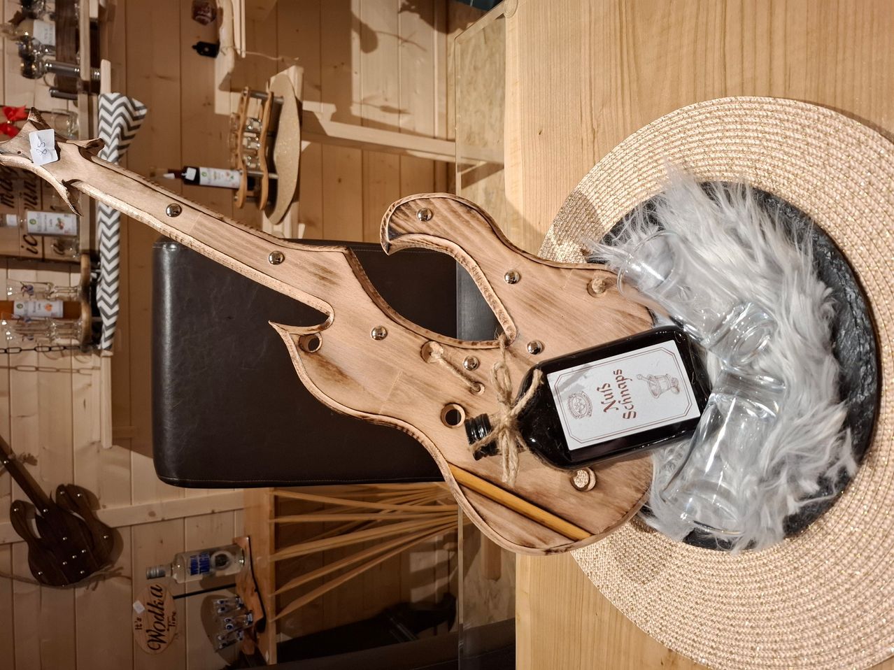 Eine hölzerne Gitarre steht auf einem Stuhl in einem Raum mit Holzwänden. Sie hält eine Flasche Schnaps, um sie herum befindet sich ein Hut und Gläser. Eine Flasche Alkohol steht ebenfalls auf dem Boden in der Nähe des Stuhls.