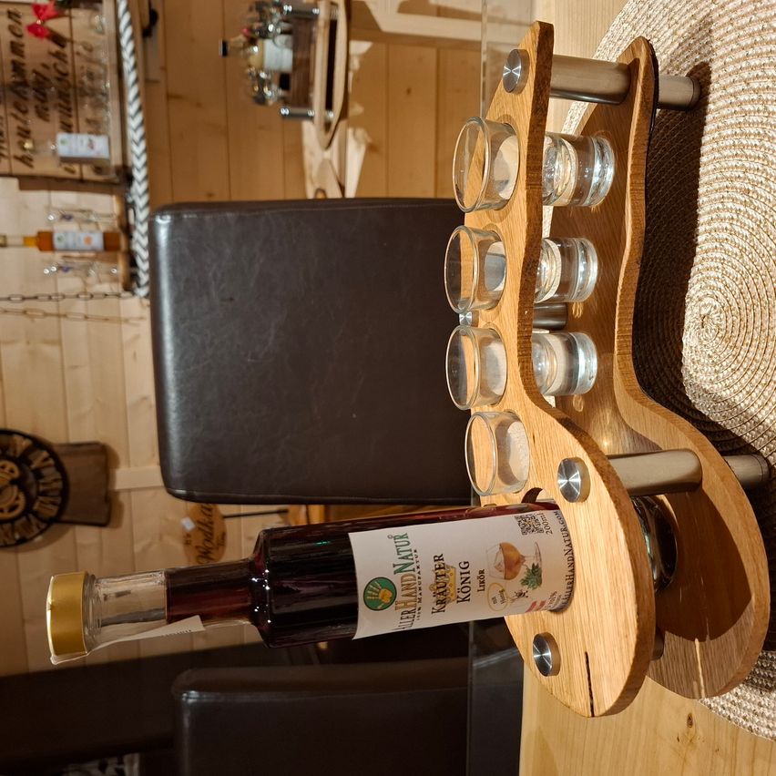 Eine Flasche Likör steht auf einem Holzhalter mit vier Gläsern darüber, in einem Raum mit Holzwänden und einem Lederstuhl.