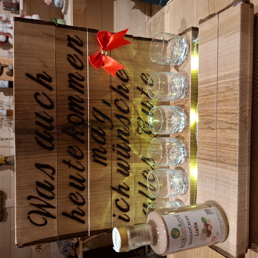 Ein Holzschild mit einer roten Schleife und dem Spruch 'Was auch heute morgen ich wünsche' ist angebracht. Vier leere Gläser stehen neben dem Schild, und eine Flasche Haselnusscreme lehnt daran.