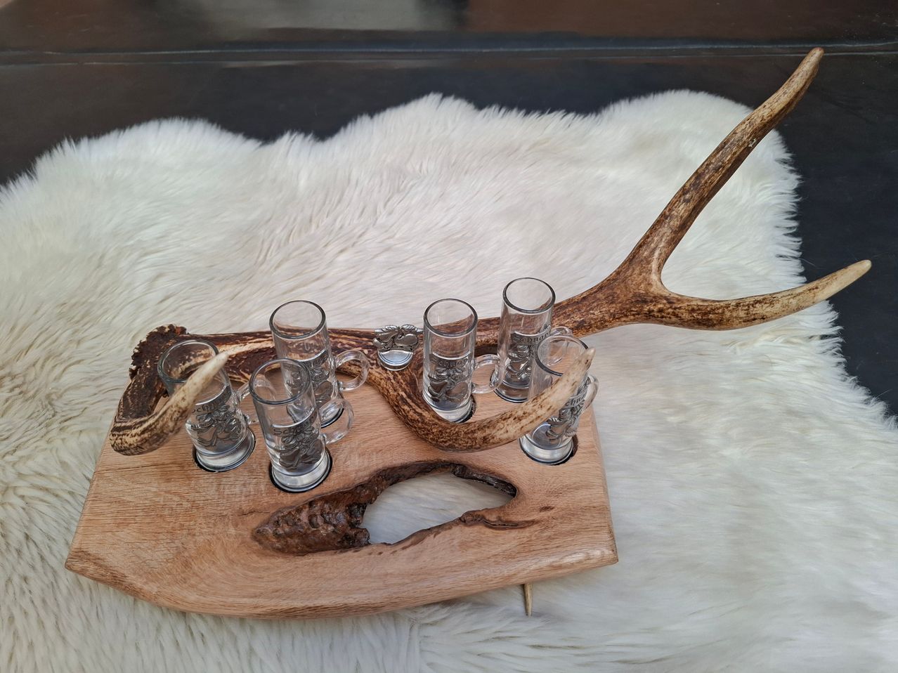 Ein Holztablett mit Hirschgeweih, mit sechs klaren Shotgläsern, steht auf einer weißen Felloberfläche.
