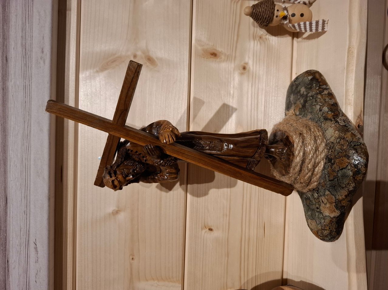 Ein hölzernes Kruzifix mit einem Mann, der ein Kreuz trägt, ist an einer Holzwand befestigt. Ein Seil ist an das Kreuz gebunden.