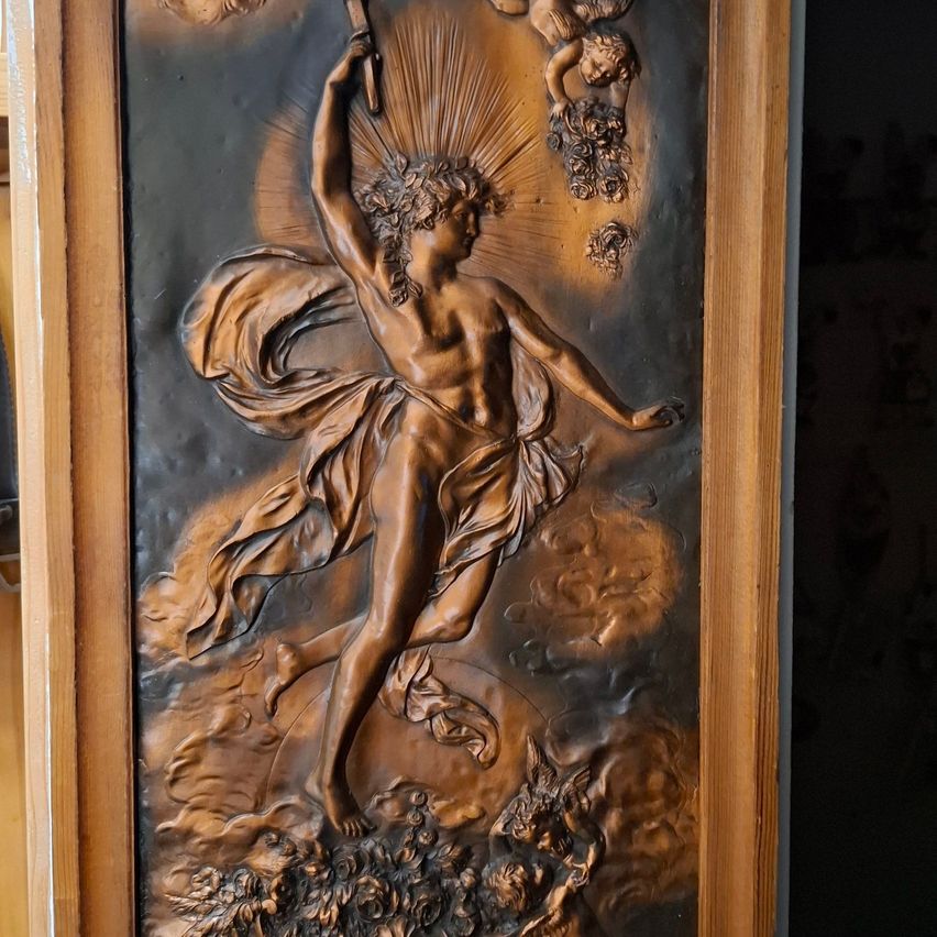 Ein Bronzerelief, das eine Figur eines Mannes zeigt, der einen Stab mit Lichtstrahlen in der Hand hält, die von ihm ausgehen, und über einem Blumenarrangement schwebt. Das Bildnis ist in einem Holzrahmen eingefasst.