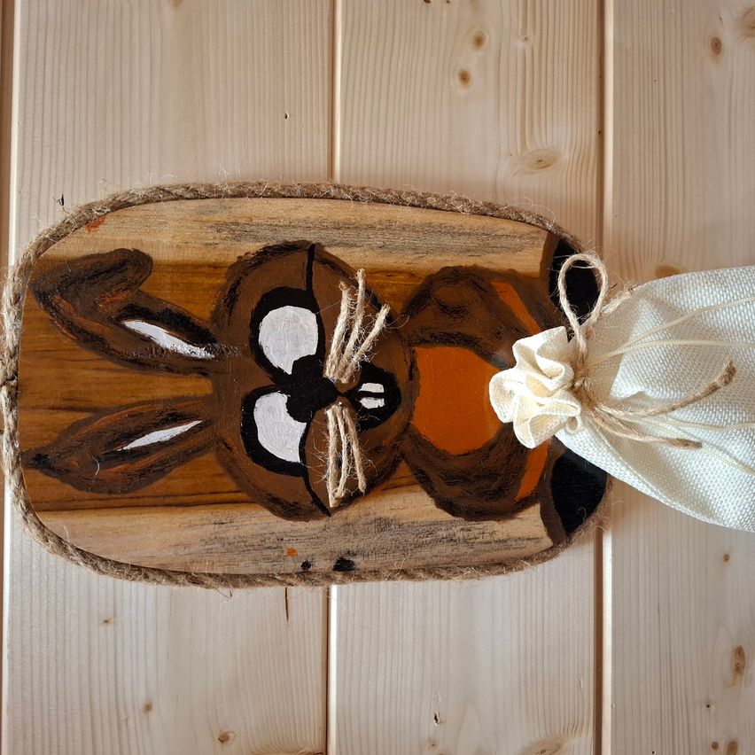 Eine Holzplatte mit einem gemalten Hasen, mit einem Seil und einem weißen Band befestigt, hängt an einer Holzwand. Der Hase hat braune und orangefarbene Farben mit schwarzen Augen und weißen Flecken.