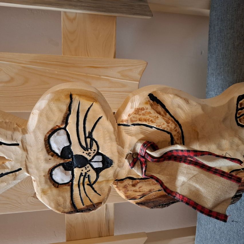 Eine Holzskulptur einer Maus mit detaillierten schwarzen und weißen Gesichtszügen ist an einer Holzkonstruktion befestigt, mit einem rot-schwarz karierten Band darum.