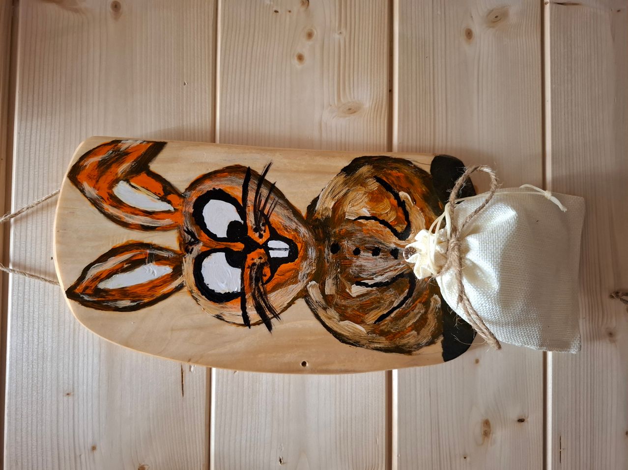 Ein Holzbrett mit einem gemalten Hasendesign darauf ist an einer Holzwand montiert. Der Hase hat orange und schwarze Farben. Es hat einen weißen Stoffbeutel, der mit einem Seil daran befestigt ist.