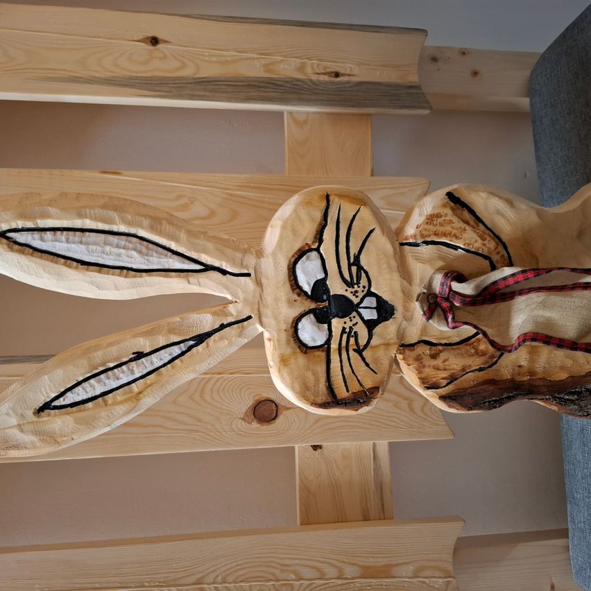 Eine Holzskulptur eines Hasen mit großen Ohren und einem karierten Band. Die Skulptur ist aus einem Stück Holz geschnitzt.