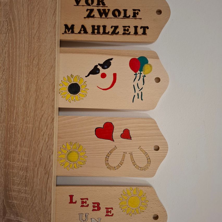 Four wooden signs with different decorations are mounted on a wall. The top sign reads 'Vor Zwolf Mahlzeit'. The middle sign has a clown, balloons, and a sunflower. The third sign features two red hearts and horseshoes. The bottom sign reads 'Lebe Unbe'.