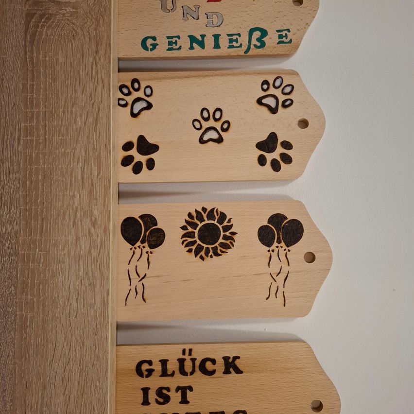 Four wooden signs are mounted on a wall. The top sign says 'und genieße'. The middle left sign has a paw print, and the middle right sign has two balloons. The bottom sign says 'Gluck ist'.
