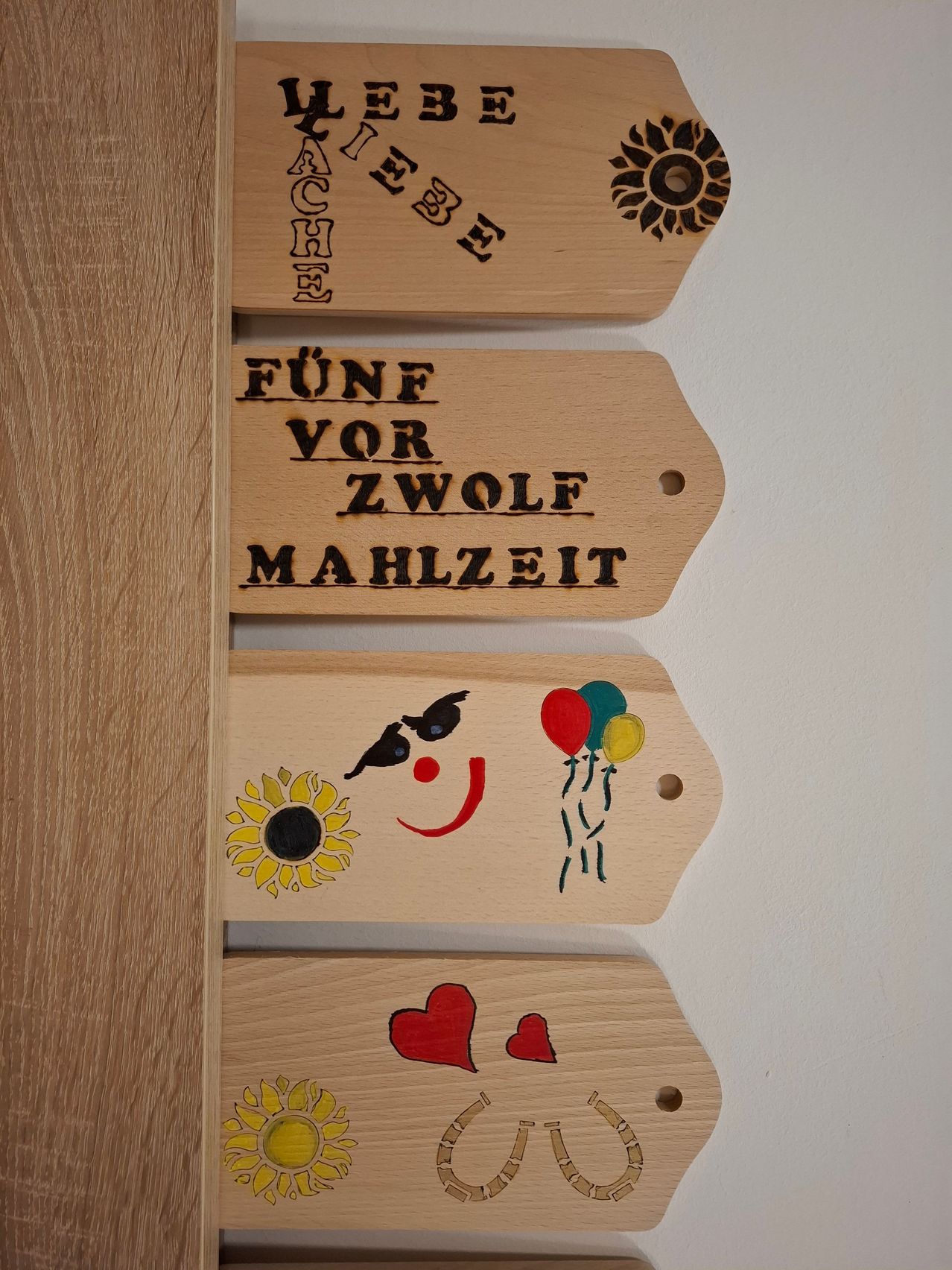 A row of wooden tags on a white wall. The top tag reads 'Free' in cursive. Below it, there are three more tags. The middle one says 'Five before twelve mealtime'. The bottom tag has a sunflower and a smiley face.