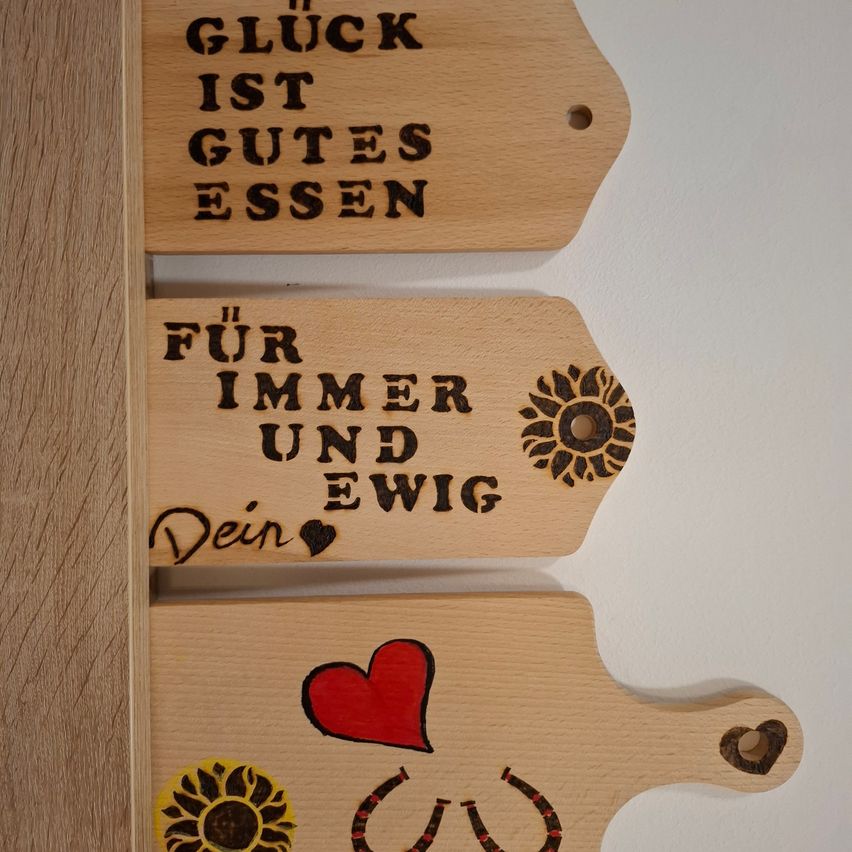Three wooden plaques on a wall with German text. The top plaque reads 'Gluck ist gutes essen', the middle has 'Fur immer und ewig Dein' with a heart, and the bottom has a red heart and horseshoe.