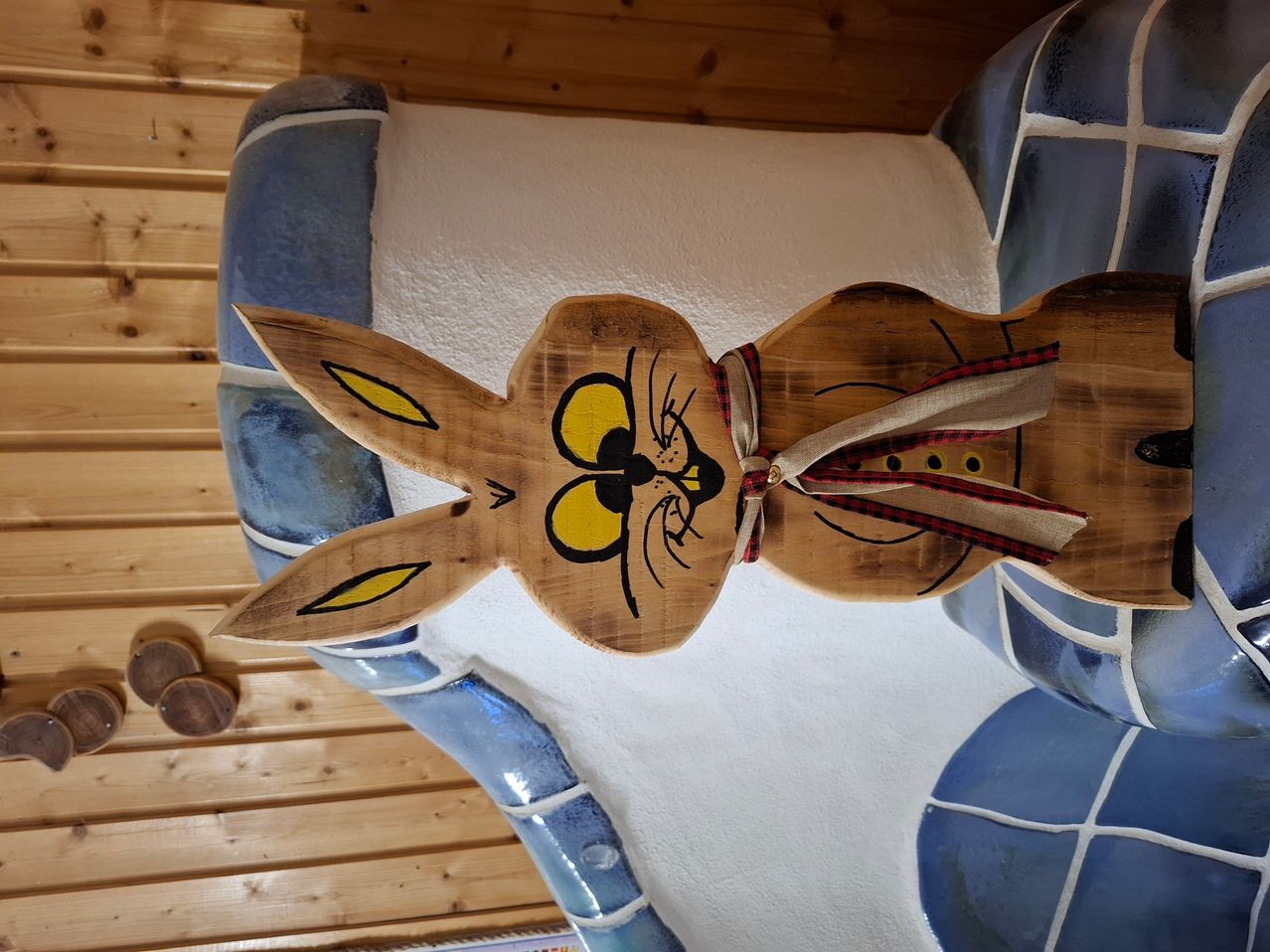 A wooden carved rabbit with yellow eyes and a red and black ribbon around its neck, mounted on a white tiled wall with blue accents.