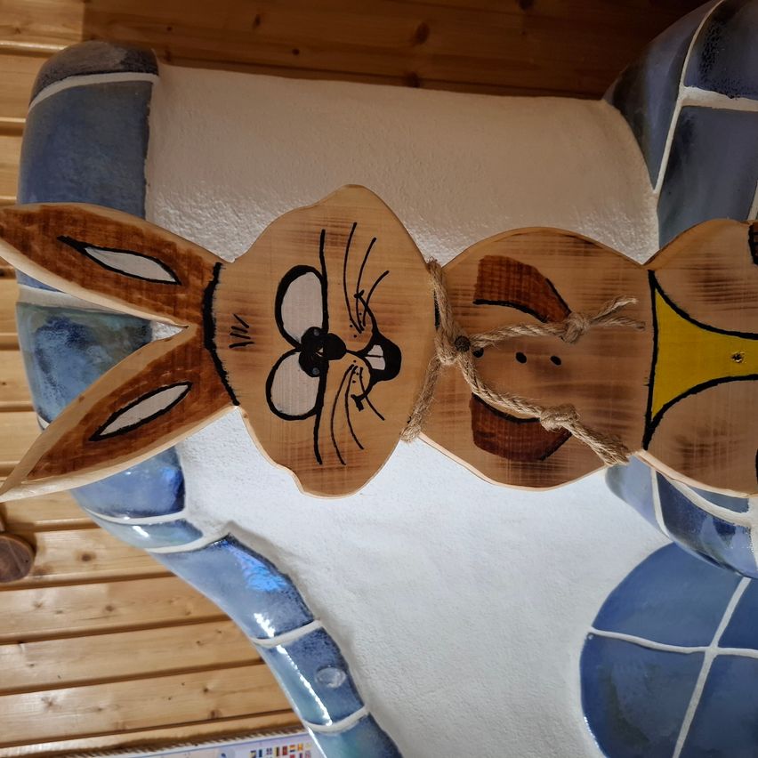 A wooden rabbit decoration hangs on a tiled wall. It has a yellow shirt and a rope around its neck. The background includes wooden paneling and tiles.