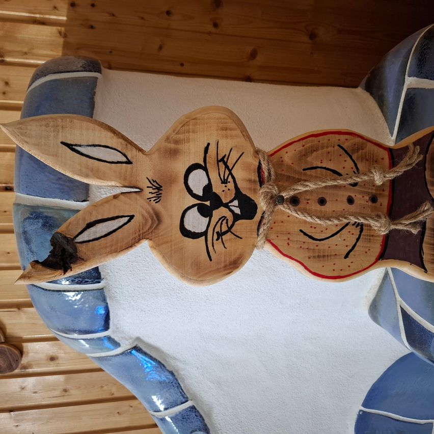 A wooden rabbit sculpture is mounted on a white surface, with blue mosaic tiles below. The rabbit has a rope tied around its neck.