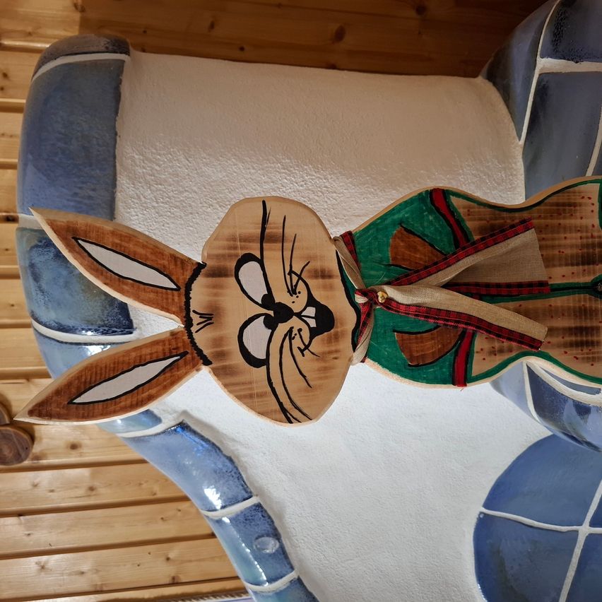 A wooden rabbit figurine is mounted on a blue and white patterned wall, against a wooden background. The rabbit has large ears, a brown body, and a green and red checkered scarf.