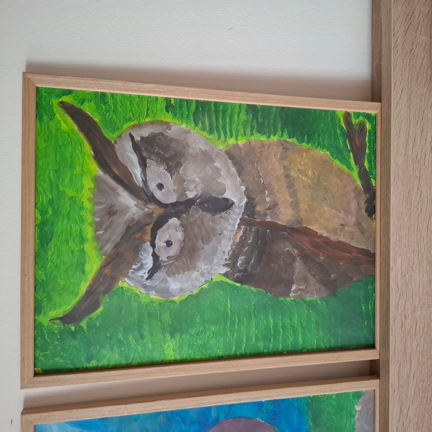 Ein gerahmtes Gemälde eines auf dem Gras liegenden Uhus. Der Uhu hat zwei Augen und ausgebreitete Flügel. Das Gemälde ist in grünen und braunen Farben gehalten.