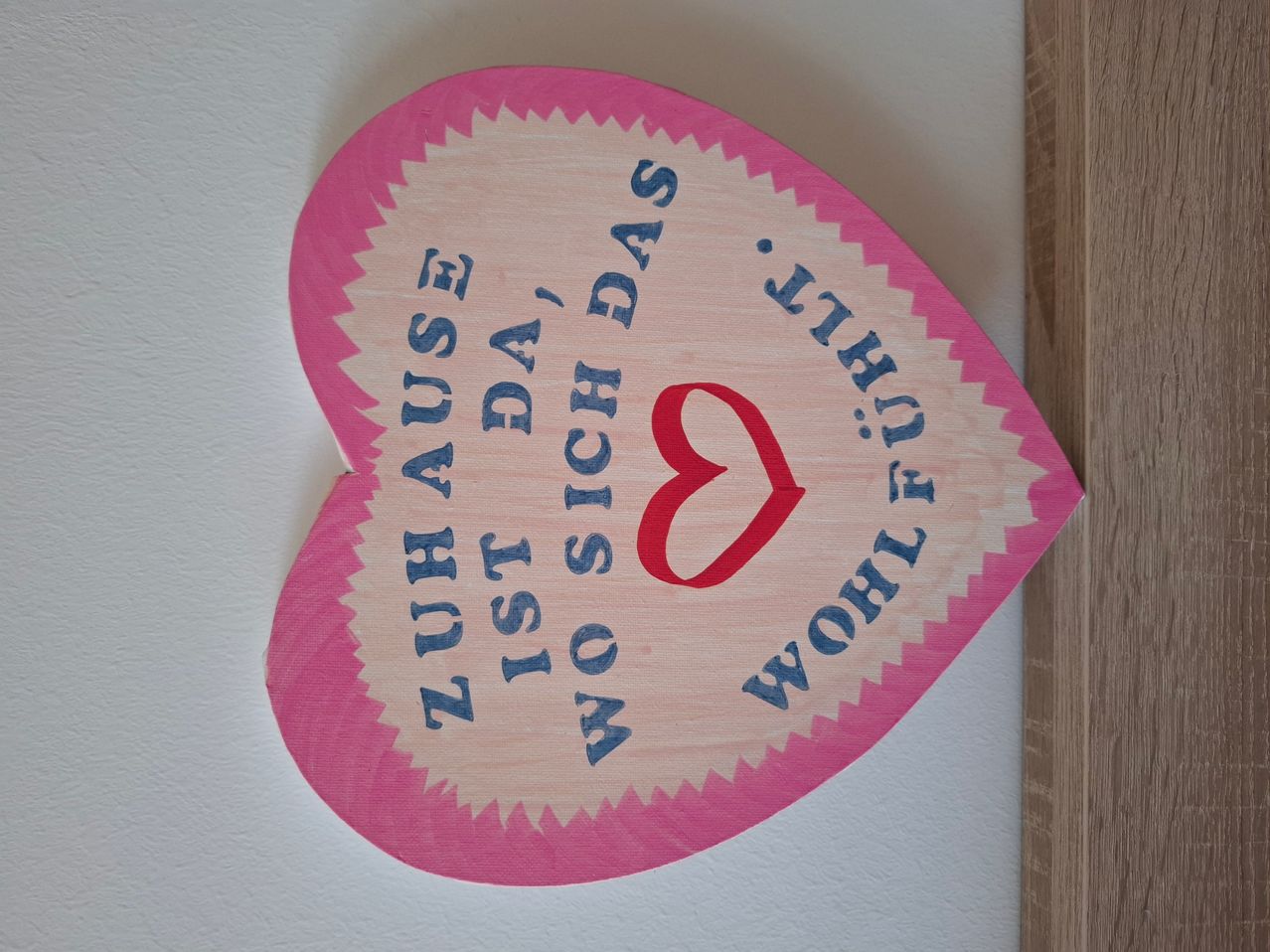 Ein herzförmiges Holzschild mit der Aufschrift 'Weil du mein Alles bist' ist an einer weißen Wand angebracht.
