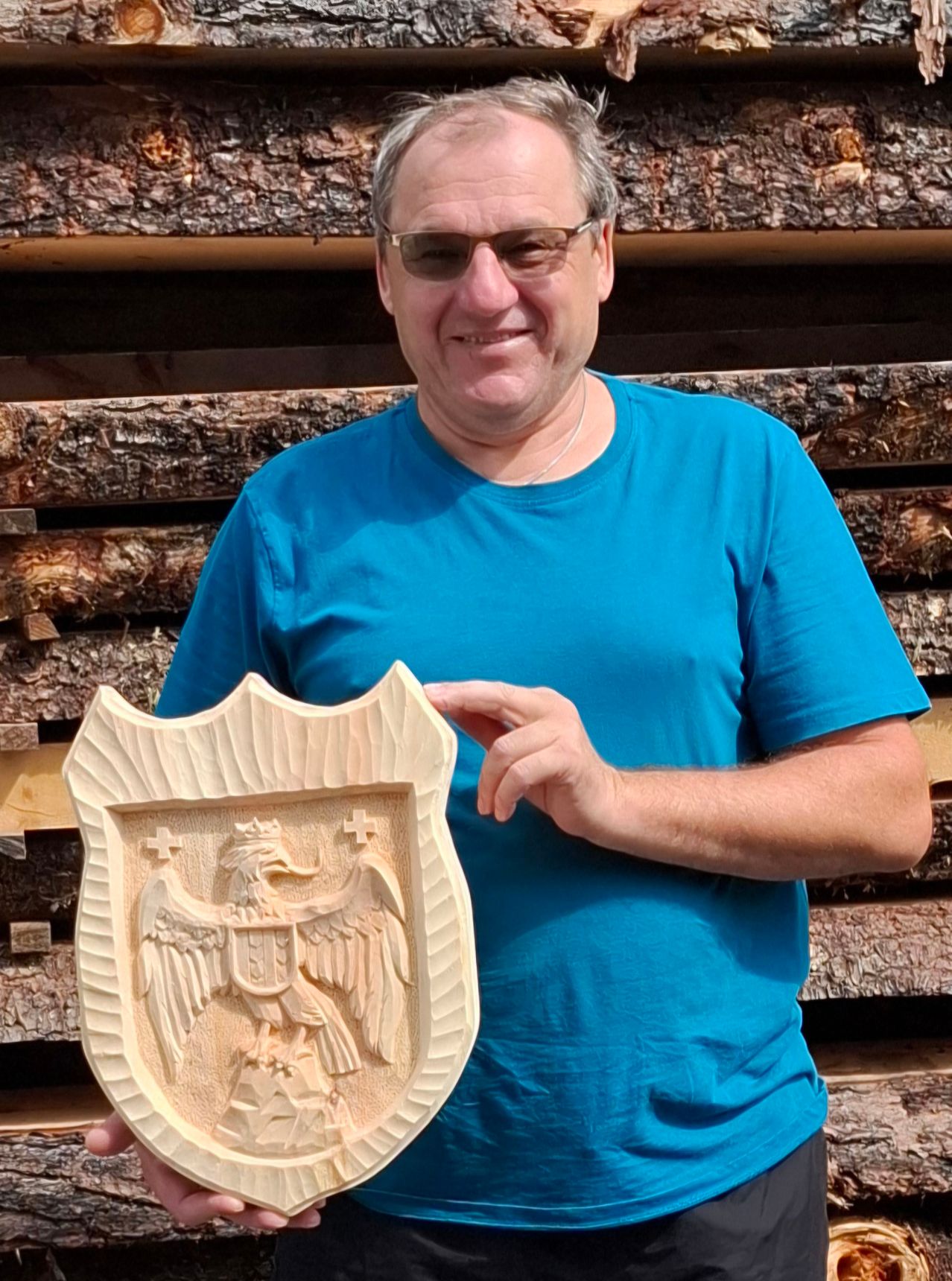 Ein Mann in einem blauen T-Shirt hält einen geschnitzten Holzschild mit einem Adlerdesign. Er steht vor einem Stapel Holzstämme.