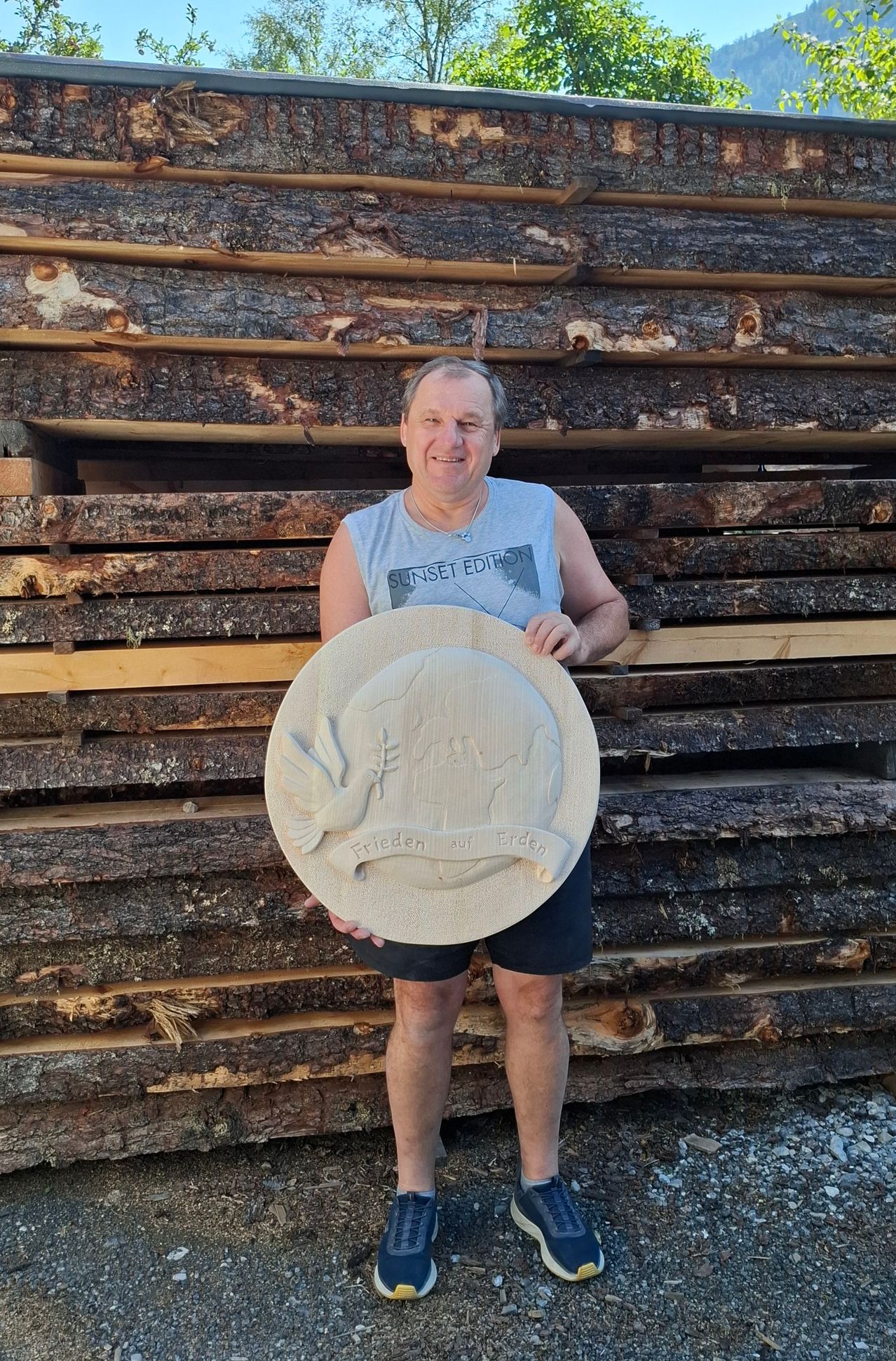 A man in a blue tank top and black shorts holds a wooden plaque with a dove and the text 'Frieden auf Erden' in front of a stack of logs.