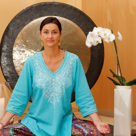 Eine Frau in einem blauen bestickten Oberteil und einem farbenfrohen Rock sitzt in einer Lotusposition vor einem großen Gong, mit einer weißen Orchidee in einem Vasen und einer Kerze in der Nähe.