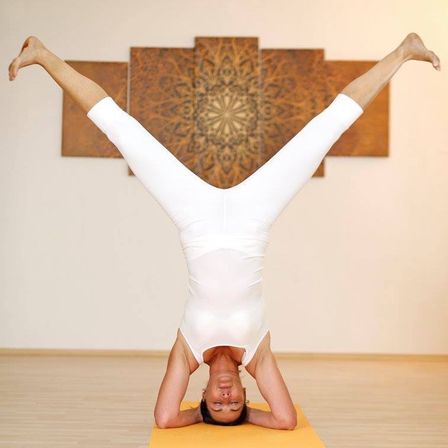 Eine Person in weißer Yoga-Kleidung vollführt einen Handstand auf einer gelben Matte in einem Raum mit Holzböden und Wandkunst.
