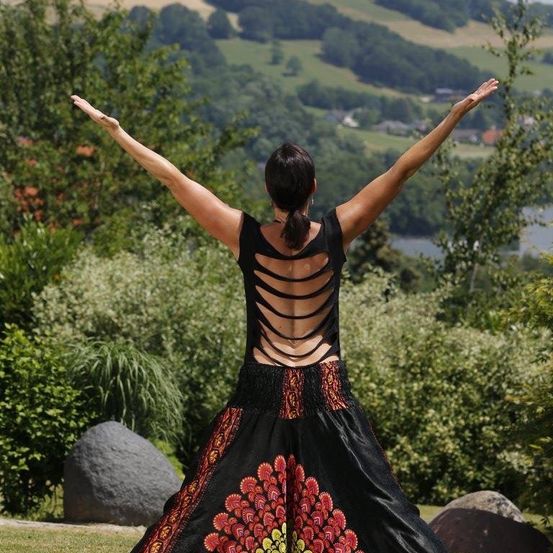 Eine Frau steht mit erhobenen Armen auf einem Hügel mit Blick auf ein Tal mit Grünflächen und einem Gewässer. Sie trägt ein schwarzes und rotes Kleid.