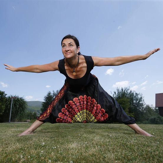 Eine Frau in einem schwarzen Oberteil und Rock praktiziert eine Yoga-Pose auf einem Rasen, Arme und Beine weit gespreizt. Hinter ihr sind Bäume und ein blauer Himmel mit Wolken zu sehen.