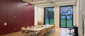 Ein leerer Konferenzraum mit einem langen Tisch, Stühlen und Glastüren, die zu einem Rasen führen. Der Raum hat eine weinrote Wand, einen Projektor und Deckenleuchten.