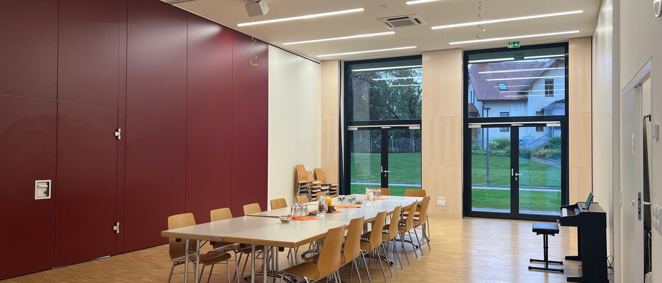 Ein leerer Konferenzraum mit einem langen Tisch, Stühlen und Glastüren, die zu einem Rasen führen. Der Raum hat eine weinrote Wand, einen Projektor und Deckenleuchten.