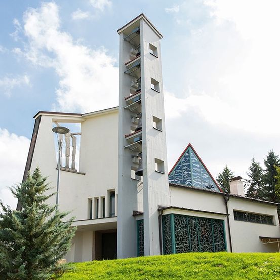 Eine moderne weiße Kirche mit einem hohen Glockenturm und einem dreieckigen Glasdach steht auf einem Rasenhügel unter einem blauen Himmel mit Wolken.