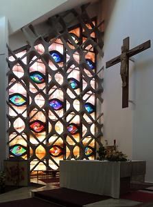 Ein Innenansicht einer Kirche mit einem großen Buntglasfenster, das mehrere farbige Augen darstellt. Ein Holzkreuz ist an der Wand über dem Altar angebracht, der mit einem weißen Tuch bedeckt und mit Blumen geschmückt ist.