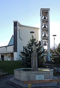 Ein kleiner Brunnen mit einer Statue in der Mitte, umgeben von Tannen. Hinter dem Brunnen steht eine weiße Kirche mit einem Glockenturm. Eine Bank ist auf der linken Seite platziert.