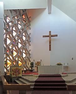 Der Innenraum einer modernen Kirche mit einem hölzernen Kreuz über dem Altar. Eine große, komplizierte Wand hinter dem Altar besteht aus Buntglasfenstern. Der Altar ist mit Blumen geschmückt und von zwei kleineren Kunstwerken flankiert.