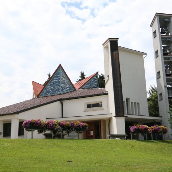 Eine moderne weiße Kirche mit einem roten Dach und einem dreieckigen Glasfenster steht auf einem grasbewachsenen Hügel. Ein Glockenturm mit Glocken befindet sich rechts.
