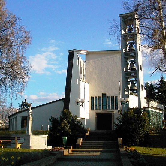 Eine weiße Kirche mit einem Glockenturm auf der rechten Seite, geschmückt mit Glocken, steht unter einem klaren blauen Himmel. Der Eingang hat einen gewölbten Eingangsbereich, flankiert von Laternenpfosten. Eine Statue steht auf einem Podest links.
