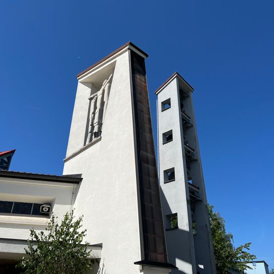 Ein hohes weißes Gebäude mit einem roten Dach hat zwei Glockentürme. Ein Turm hat ein großes Fenster und eine Skulptur einer Person. Der andere Turm hat mehrere Fenster und einen Balkon.