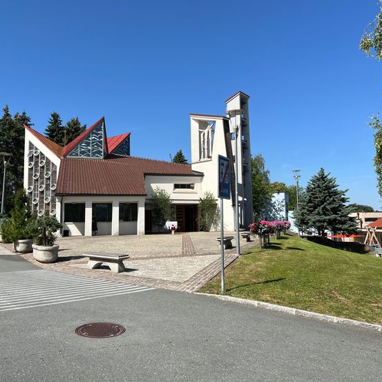 Ein modernes Gebäude mit braunem Dach und Glasfenstern, umgeben von Grün, Bänken und einem Weg.