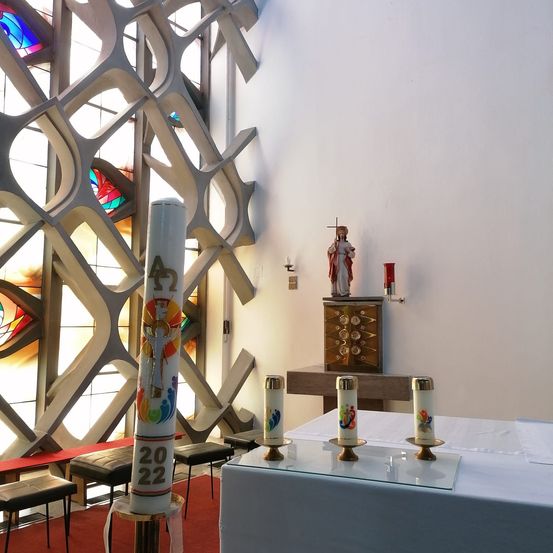 Ein Altar mit einem weißen Tuch und vier Kerzen. Dahinter hält eine Holzbox eine Statue von Jesus Christus. Eine rote und eine weiße Kerze stehen an der Wand.
