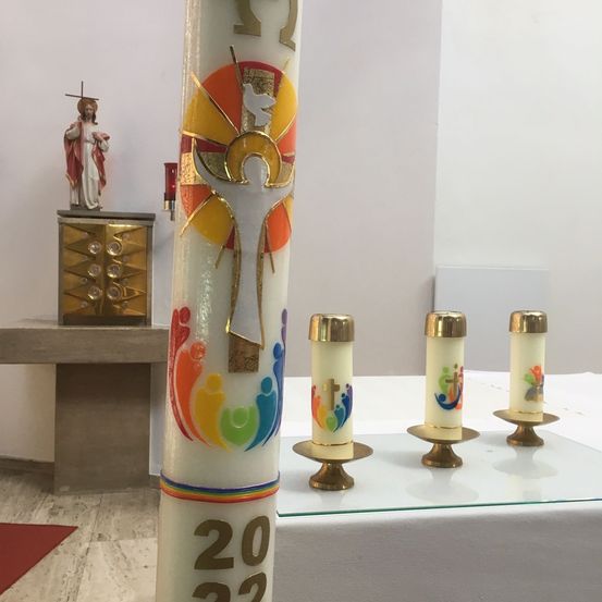 Eine weiße Kerze mit dem Jahr 2022 steht auf einem Glastisch. Dahinter ist eine rote Kerze an der Wand befestigt. Auf der linken Seite steht eine Statue von Jesus Christus auf einem Podest.