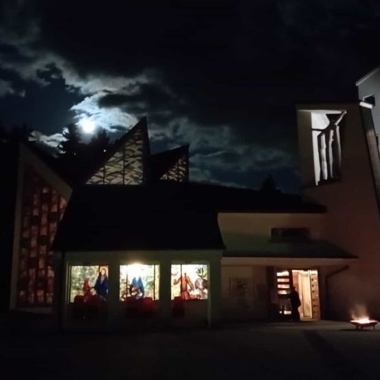 Eine Kirche ist nachts beleuchtet, mit einem Vollmond und einem Feuerkorb davor.