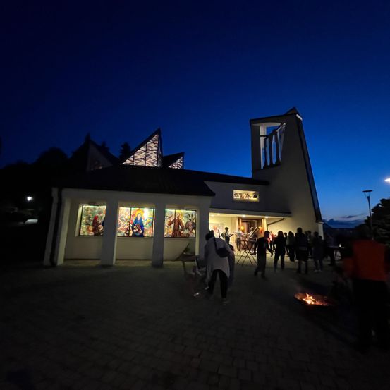 Bei Dämmerung versammeln sich einige Menschen vor einer Kapelle mit beleuchteten Buntglasfenstern. Einige stehen, während andere um ein Feuer herumsitzen.