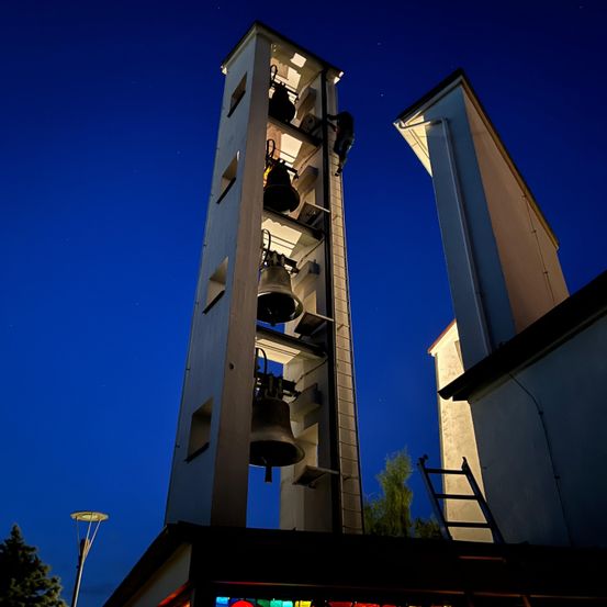 Ein hoher Glockenturm mit mehreren Glocken ist nachts beleuchtet und wirft ein warmes Licht. Eine Person klettert die Leiter hinauf, um die Spitze zu erreichen.