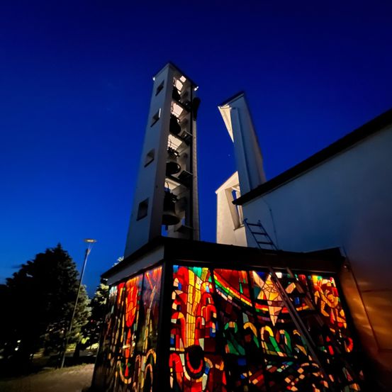 Ein hoher Turm mit Glocken steht hinter einem Gebäude mit einem farbenfrohen Buntglasfenster. Der Turm ist beleuchtet und das Gebäude im Hintergrund. Die Szene wird vom blauen Himmel bei Dämmerung beleuchtet.