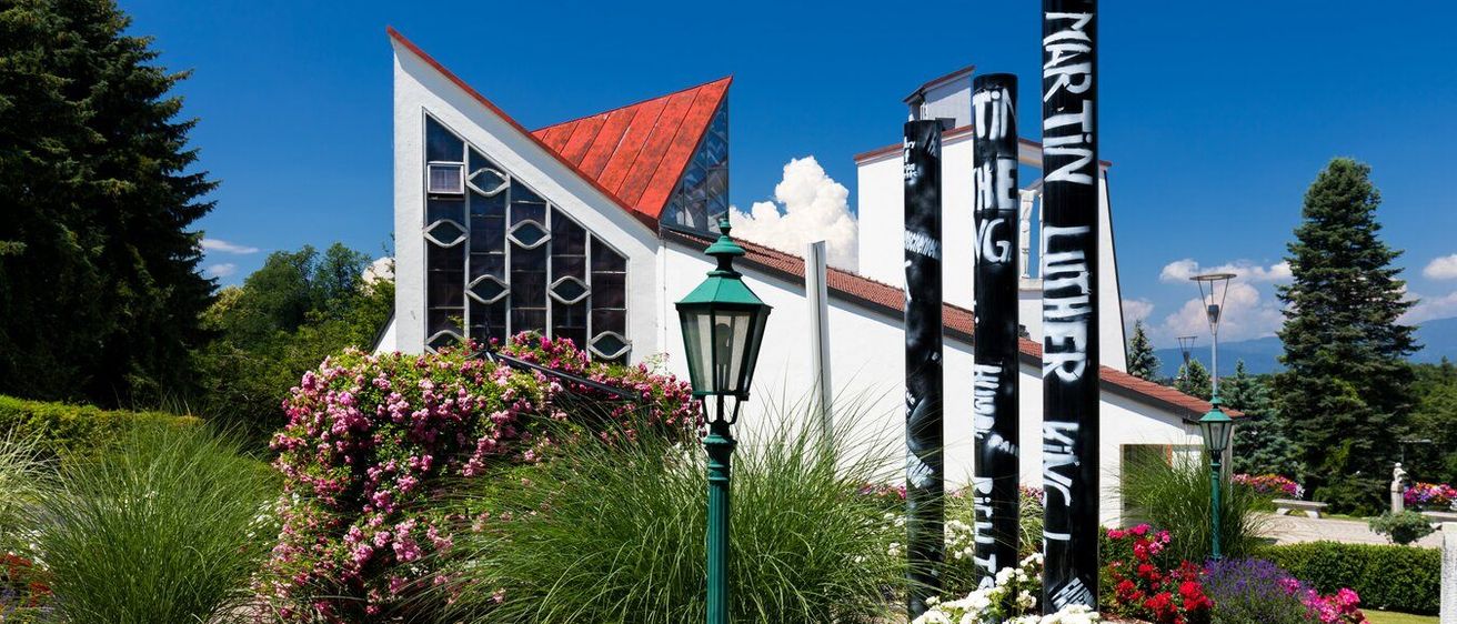 Ein Garten mit einem weißen Rosenbusch im Vordergrund und einem modernen Gebäude im Hintergrund mit einem roten Dach.