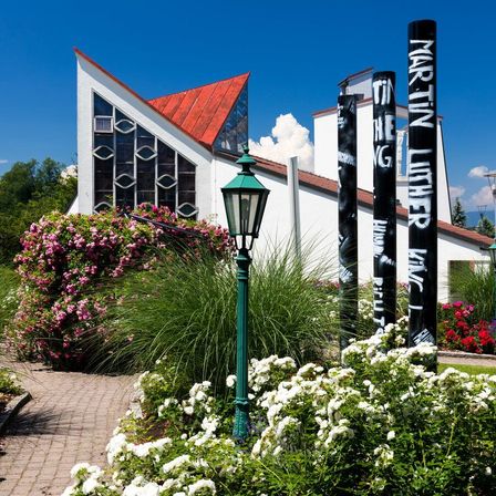 Ein modernes weißes Gebäude mit rotem Dach und großen Fenstern steht hinter einem Garten mit rosa und weißen Blumen. Zwei schwarze Pfosten mit weißem Text stehen neben einer grünen Laterne.