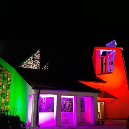 Ein modernes Gebäude mit geometrischem Design ist nachts mit roten, grünen und lila Lichtern beleuchtet. Der Eingang hat ein Glasfenster und eine Bank.