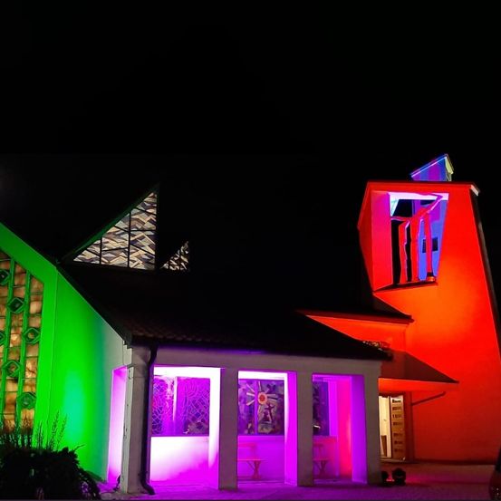 Ein modernes Gebäude mit geometrischem Design ist nachts mit roten, grünen und lila Lichtern beleuchtet. Der Eingang hat ein Glasfenster und eine Bank.