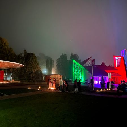 Eine Nachtszene mit einer Gruppe von Menschen, die um ein Lagerfeuer unter einem nebligen Himmel stehen. Der Bereich ist von farbigen Lichtern eines Gebäudes beleuchtet.